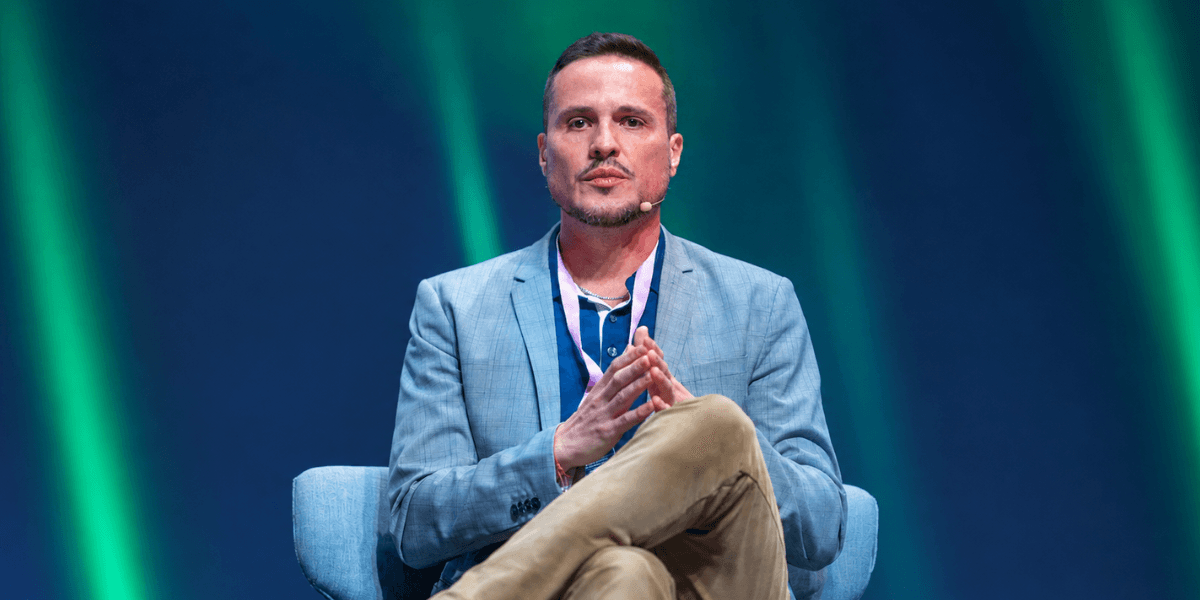 Man in a blue blazer seated on stage, hands clasped, engaged in discussion