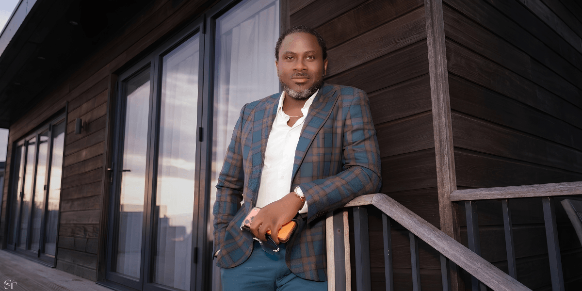 A well-dressed man poses confidently on a wooden deck with large windows behind him