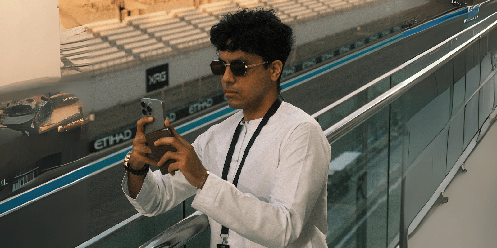 A person in sunglasses, wearing a white shirt, standing at a racetrack while looking at a mobile phone.