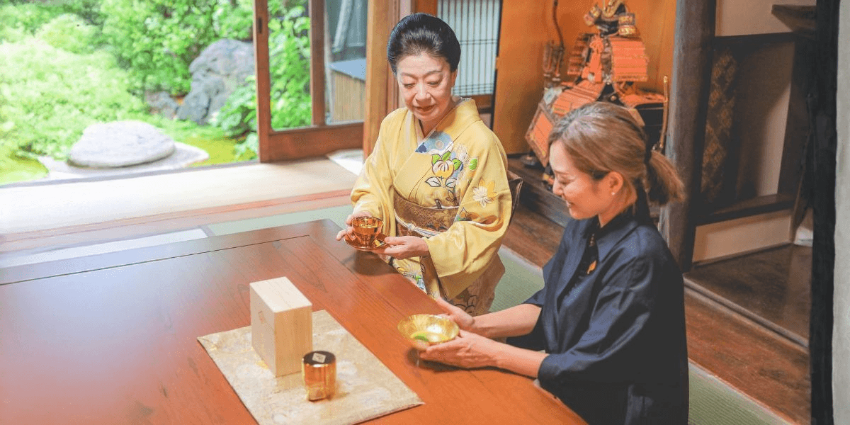 Japanese tea ceremony host in kimono serving matcha to guest at low table in traditional room.