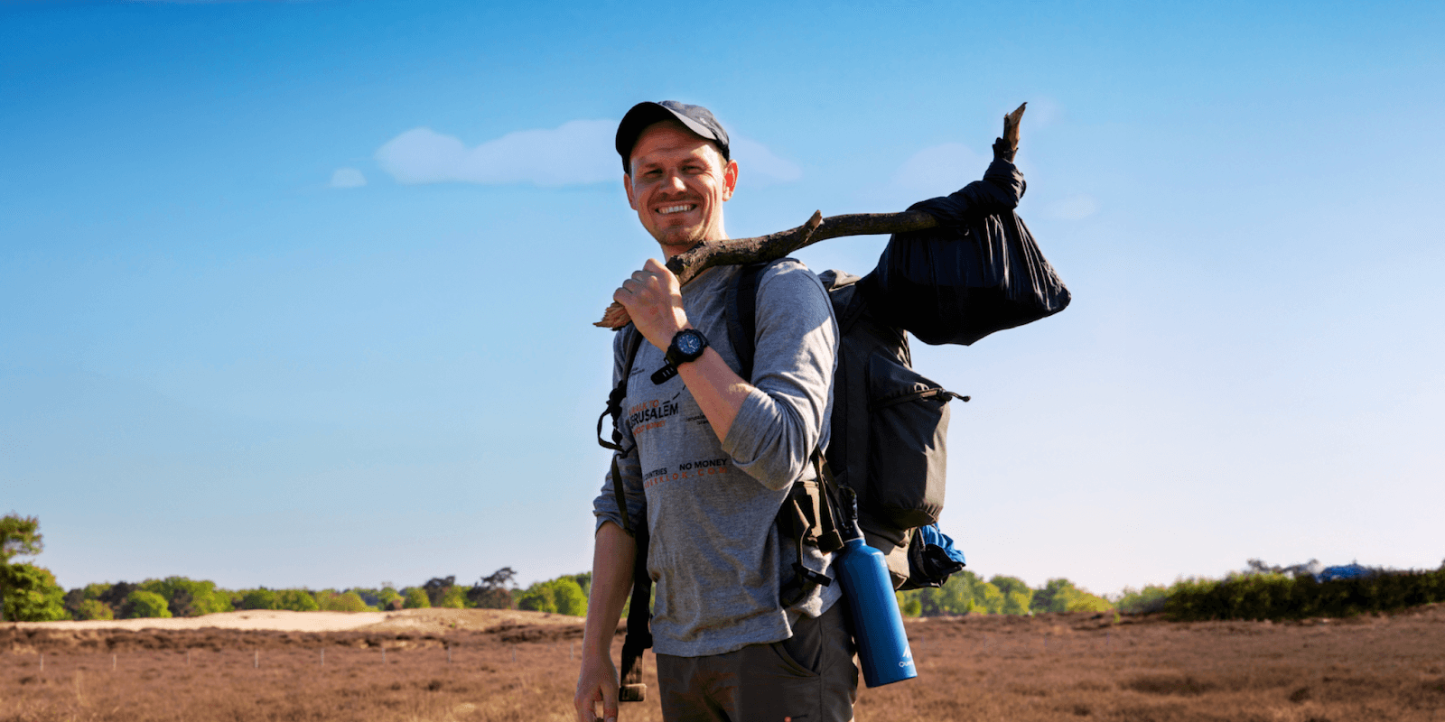 Traveler smiling while carrying a hiking backpack and a walking stick across a scenic dry landscape under a clear blue sky.