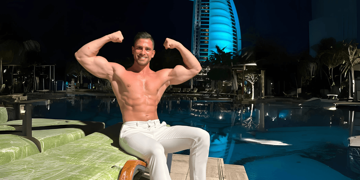 Shirtless man flexing muscles by a pool at night, with Burj Al Arab hotel illuminated in the background.