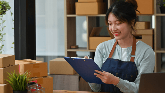 Smiling woman in an apron holds a clipboard, surrounded by cardboard boxes and a laptop