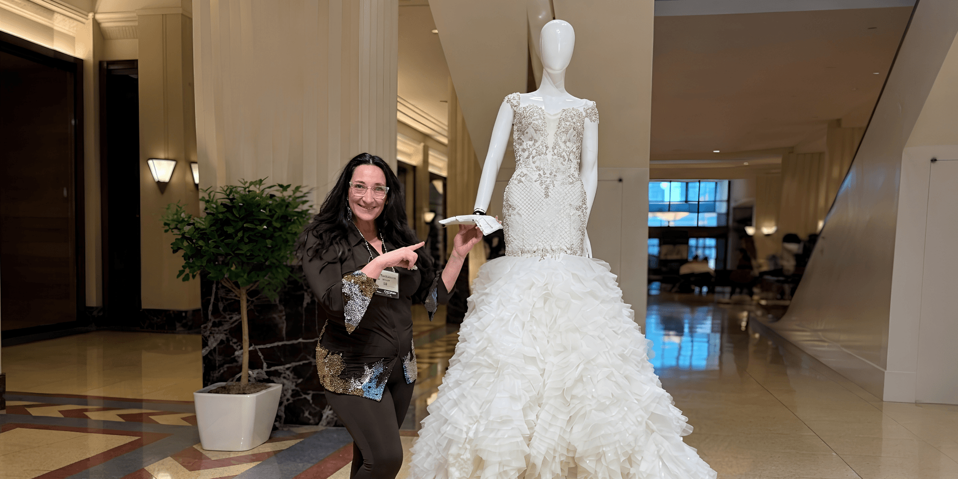 A woman poses beside a mannequin wearing an elegant ruffled wedding dress in a lobby