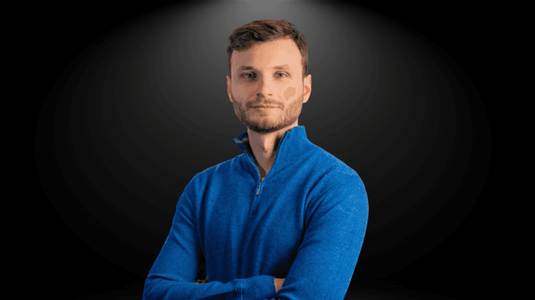 A young man wearing a blue sweater with arms crossed against a dark background