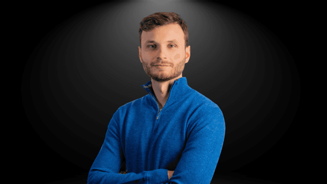 A young man wearing a blue sweater with arms crossed against a dark background