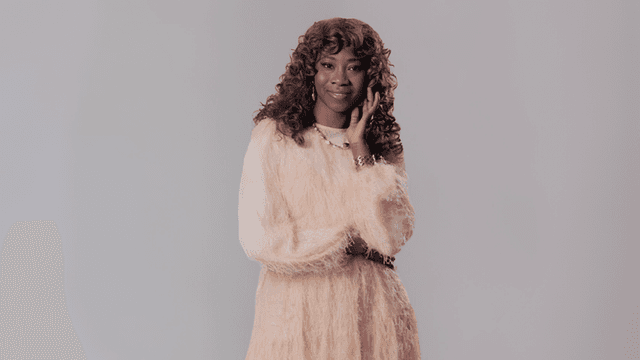 A woman with curly hair in a fluffy, light-colored dress poses elegantly against a neutral background