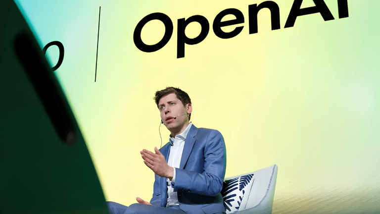 Person in a blue suit speaking on stage with OpenAI branding in the background