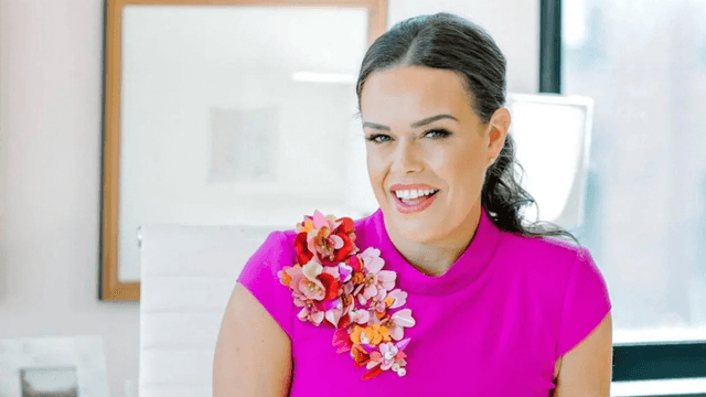Woman in a bright pink top with floral embellishments smiles confidently in an office setting