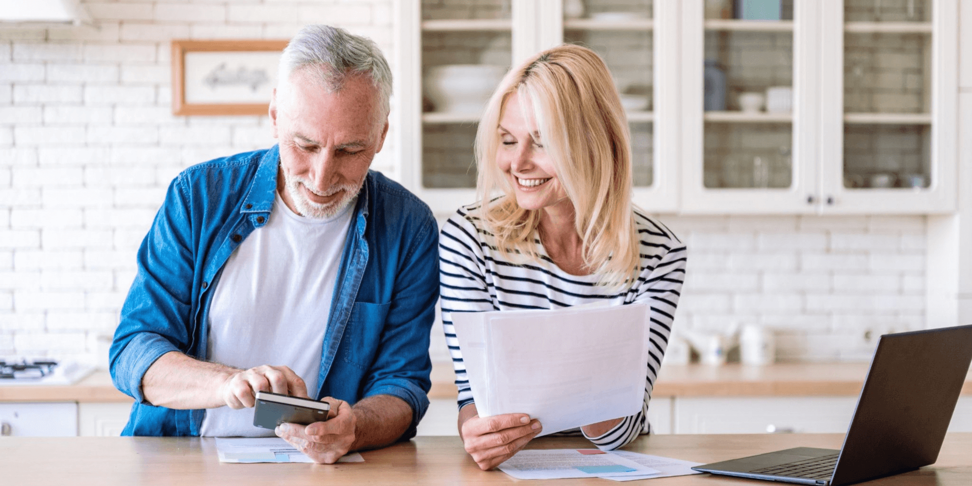Smiling man and woman discussing documents at a kitchen table with a laptop nearby