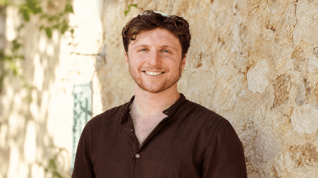 Smiling man in a brown shirt standing against a stone wall outdoors