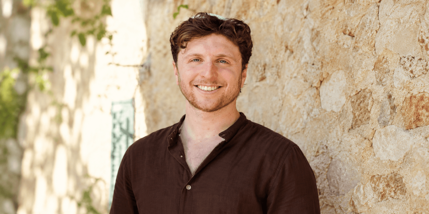 Smiling man in a brown shirt standing against a stone wall outdoors