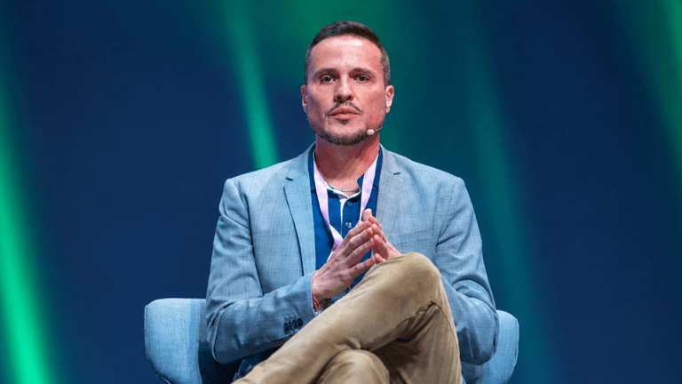 Man in a blue blazer seated on stage, hands clasped, engaged in discussion