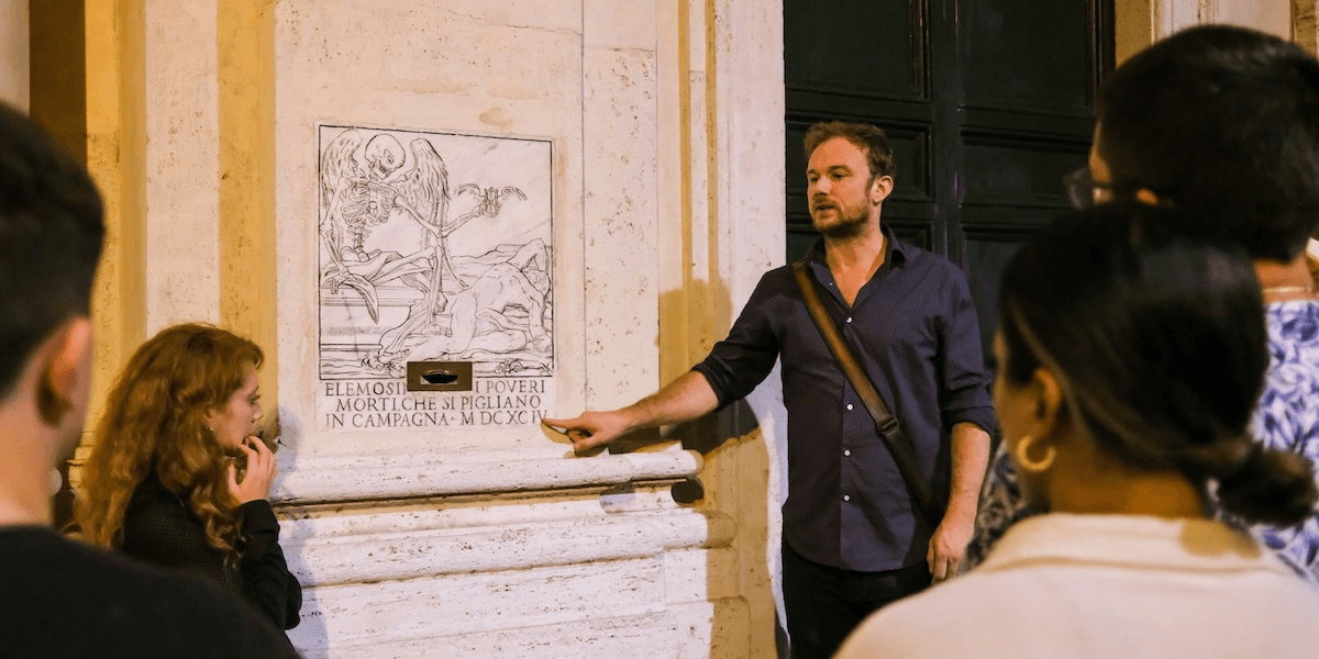 A tour guide explaining Rome's dark history to a small group, standing beside an ancient plaque depicting a skeleton.