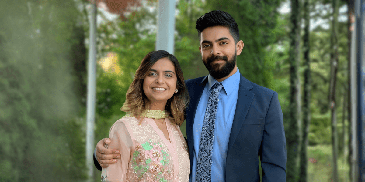 A couple posing outdoors, smiling confidently, with the woman in a traditional dress and the man in a suit.