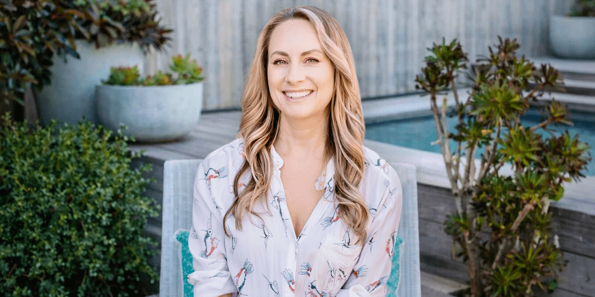 Smiling blonde woman seated outdoors near a pool and garden, wearing a patterned blouse.