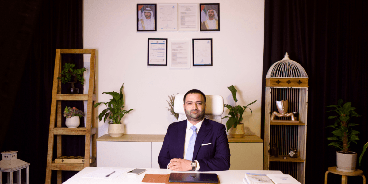 Businessman seated at a modern office desk with framed certificates and plants in the background.