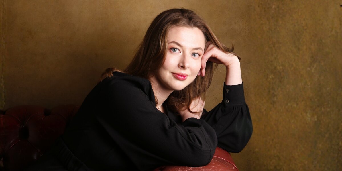 Woman in black blouse with light brown hair leans on a leather sofa, smiling softly. The background is a textured, warm-toned wall, creating a relaxed, elegant mood.