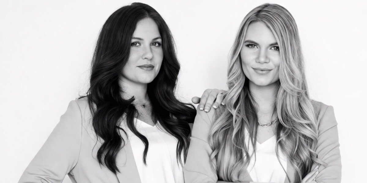 Two women posing confidently in business attire, standing side by side against a plain white background. Both have long hair and wear blazers, exuding professionalism.
