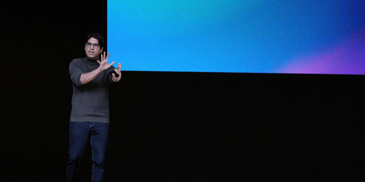 "Person giving a presentation on stage with a vibrant, abstract background, using hand gestures to explain a point, dressed in a dark sweater and glasses."