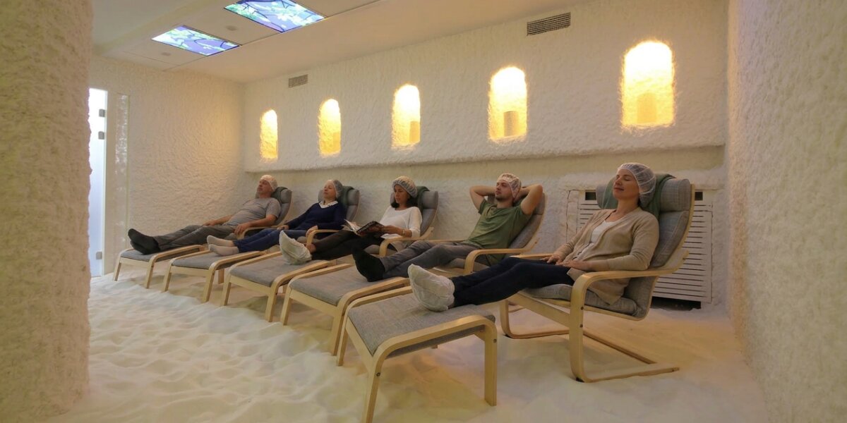 Five people in a serene salt cave lounge on wooden chairs with footrests, surrounded by salt-covered walls and floor, under soft, glowing lights.