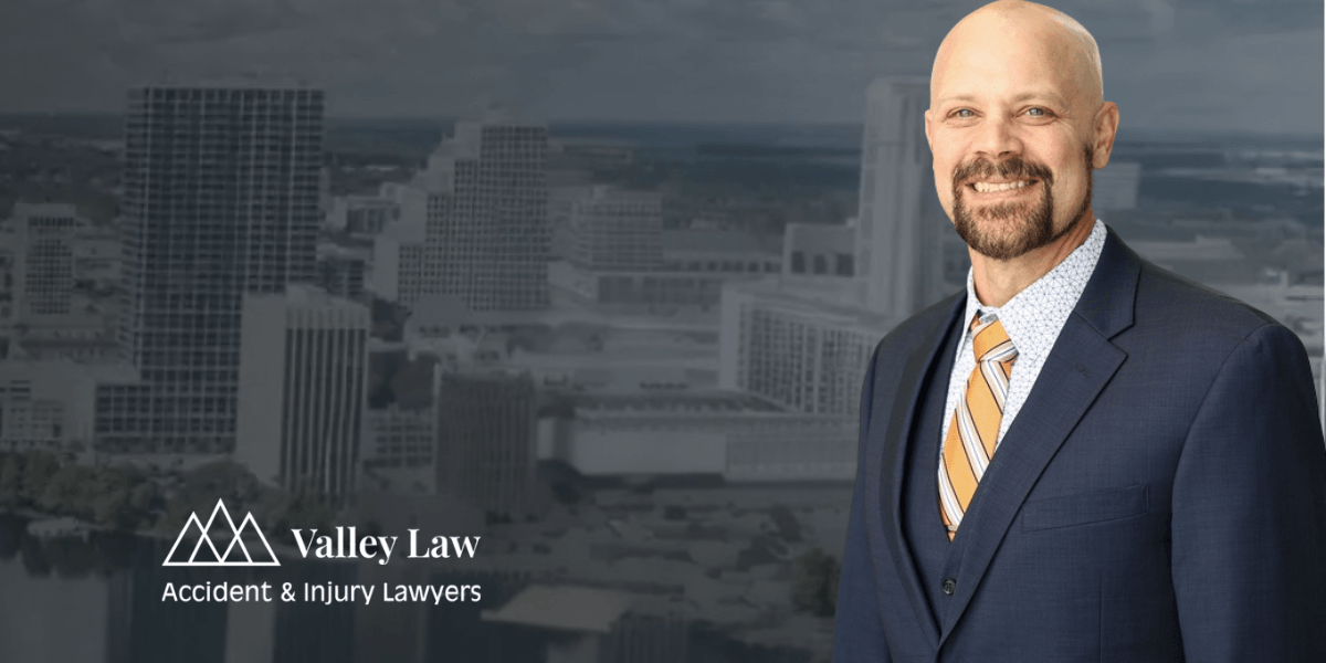 Professional portrait of a man in a suit with a patterned tie, smiling, with a city skyline background and "Valley Law" logo.
