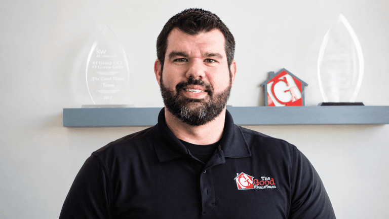 Nick Good wearing a black The Good Home Team polo with company awards displayed behind him.