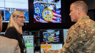 Military officer speaks with personnel inside a command center surrounded by computer monitors and U.S. military displays.