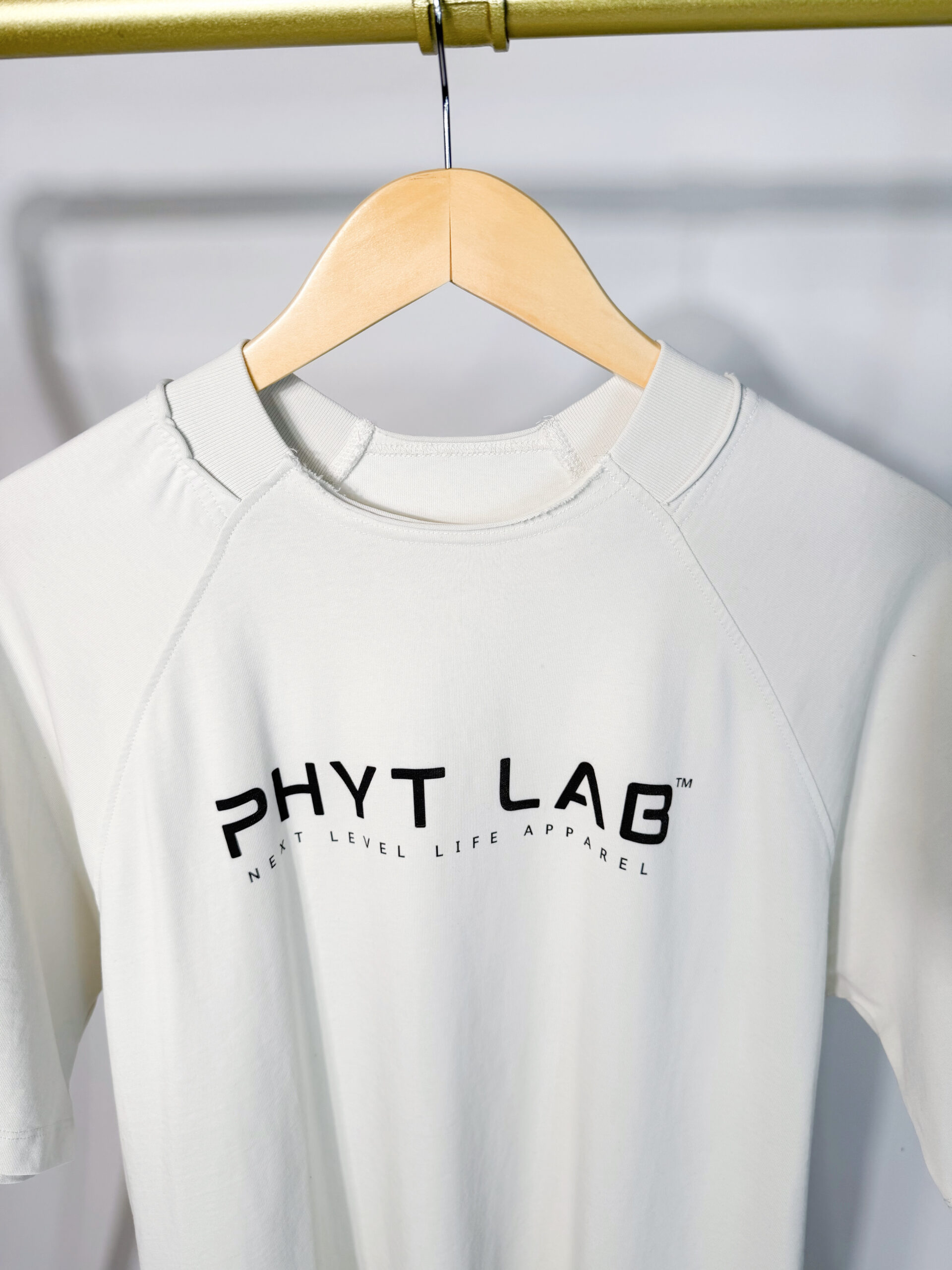 Close-up of a white t-shirt hanging on a hanger, featuring the 'PHYT LAB' logo and 'Next Level Life Apparel' text printed on the front.