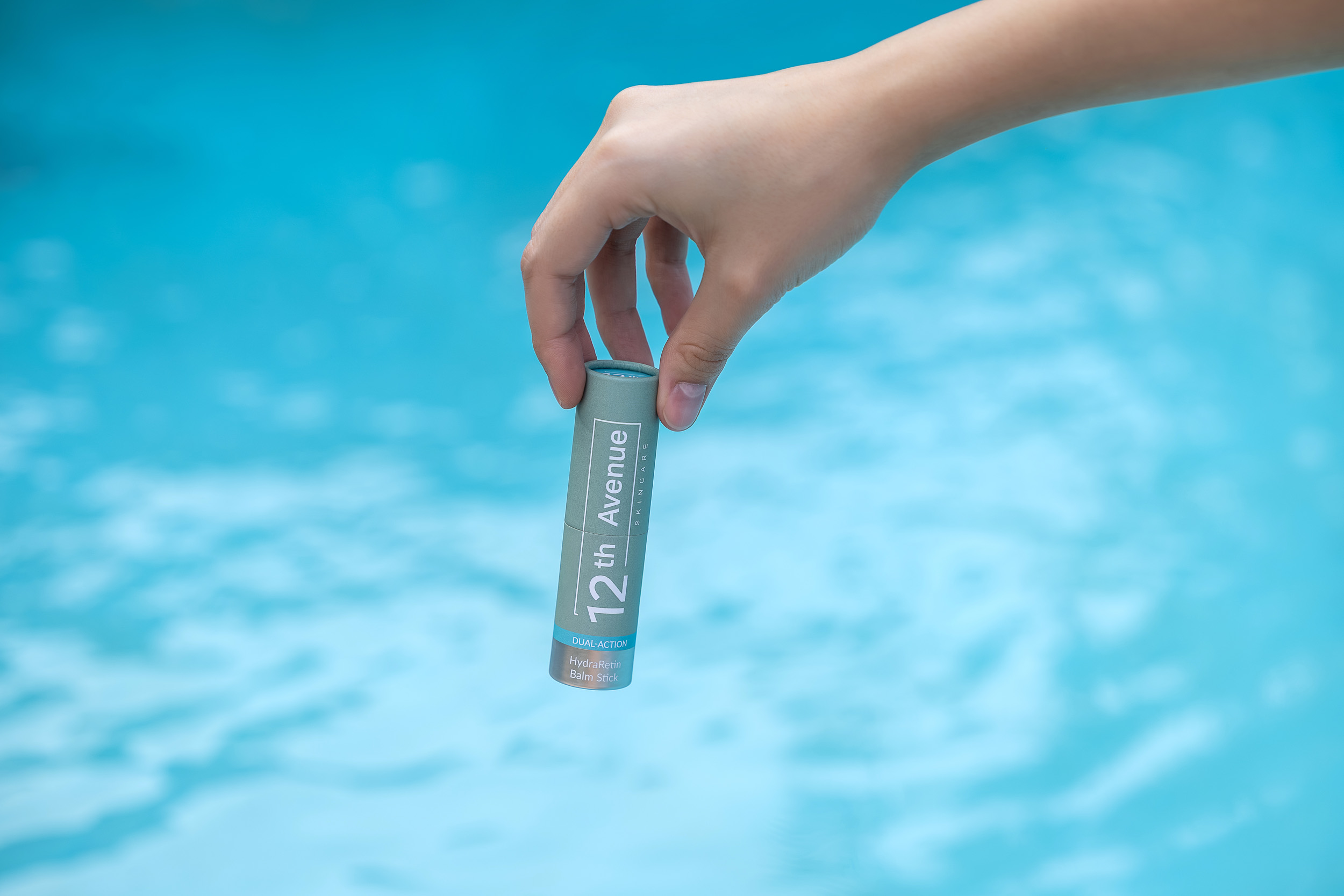 A hand holding a 12th Avenue skincare HydraRetin balm stick above a serene pool background, showcasing the product packaging and its dual-action formula for hydration and skin rejuvenation.