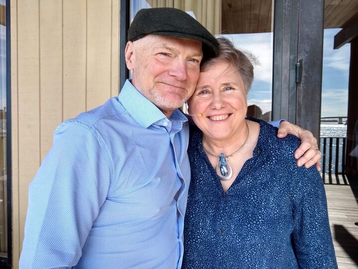A smiling couple standing close together outdoors, with the man wearing a flat cap and blue shirt, and the woman in a blue top with a necklace. They appear happy and relaxed in front of a scenic backdrop.