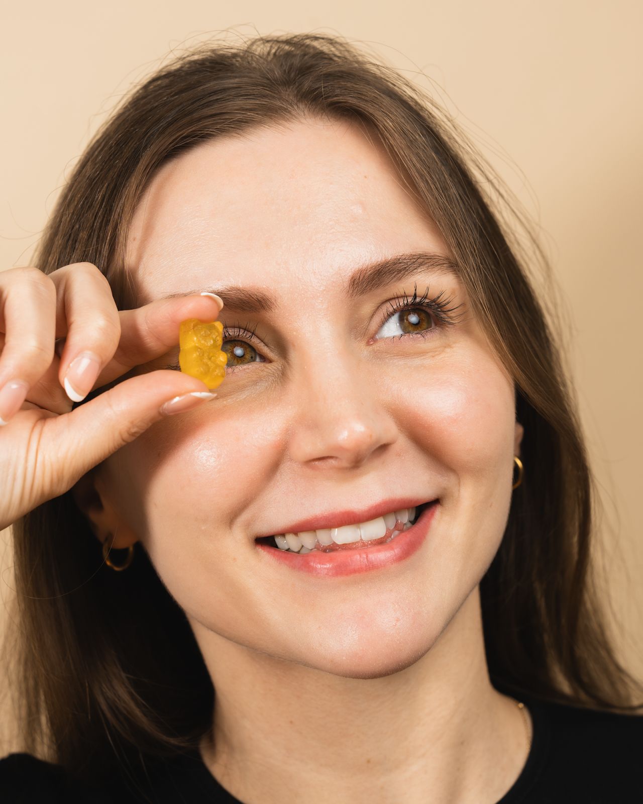 A woman smiles and holds a Vitamin C gummy in front of her eye, with a soft beige background. She is wearing a black shirt and has subtle makeup with a natural look.