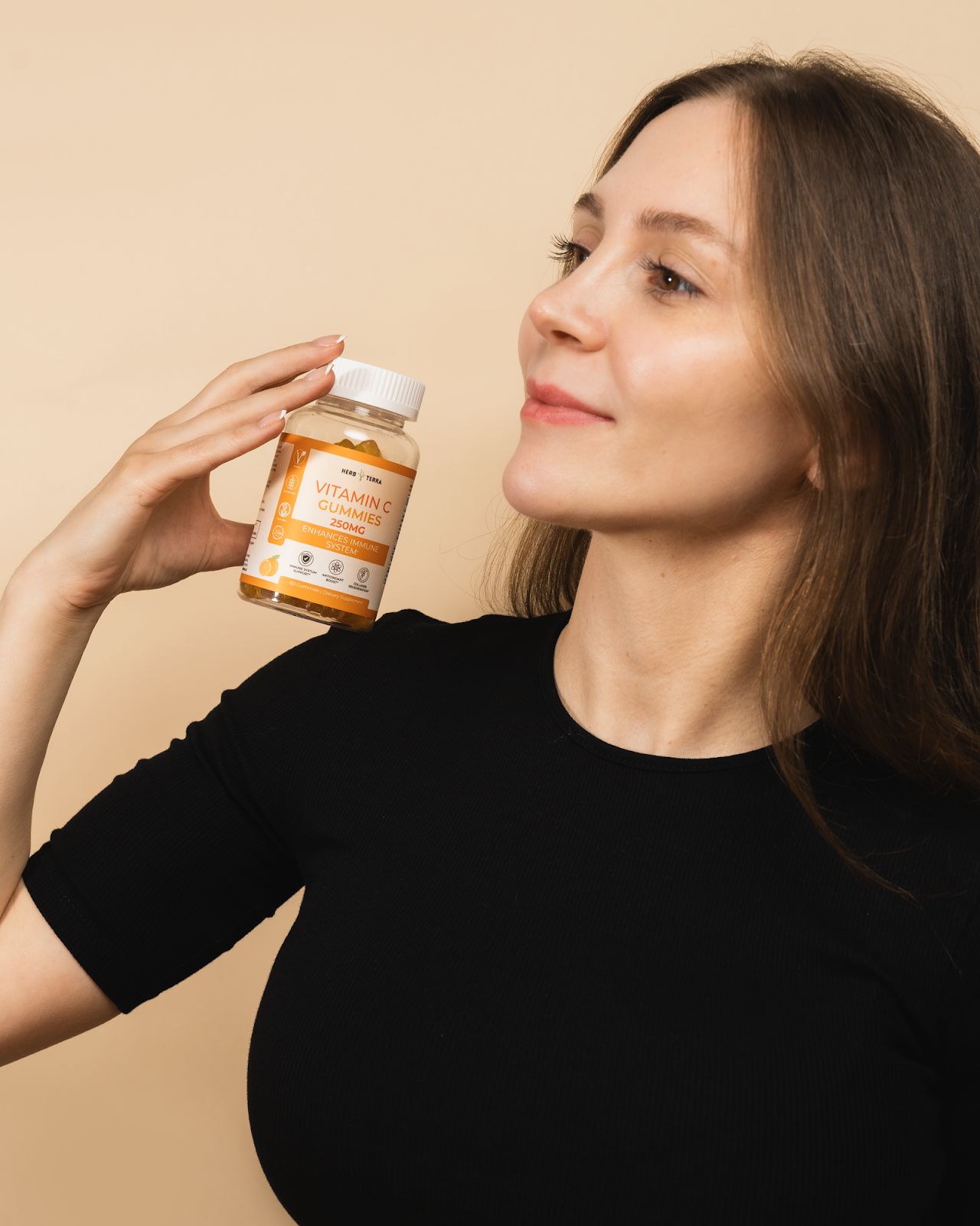 A woman in a black shirt holds a bottle of Vitamin C Gummies with a label showing '250mg, Enhances Immune Support'. The background is a soft beige, and she smiles while gazing at the bottle.