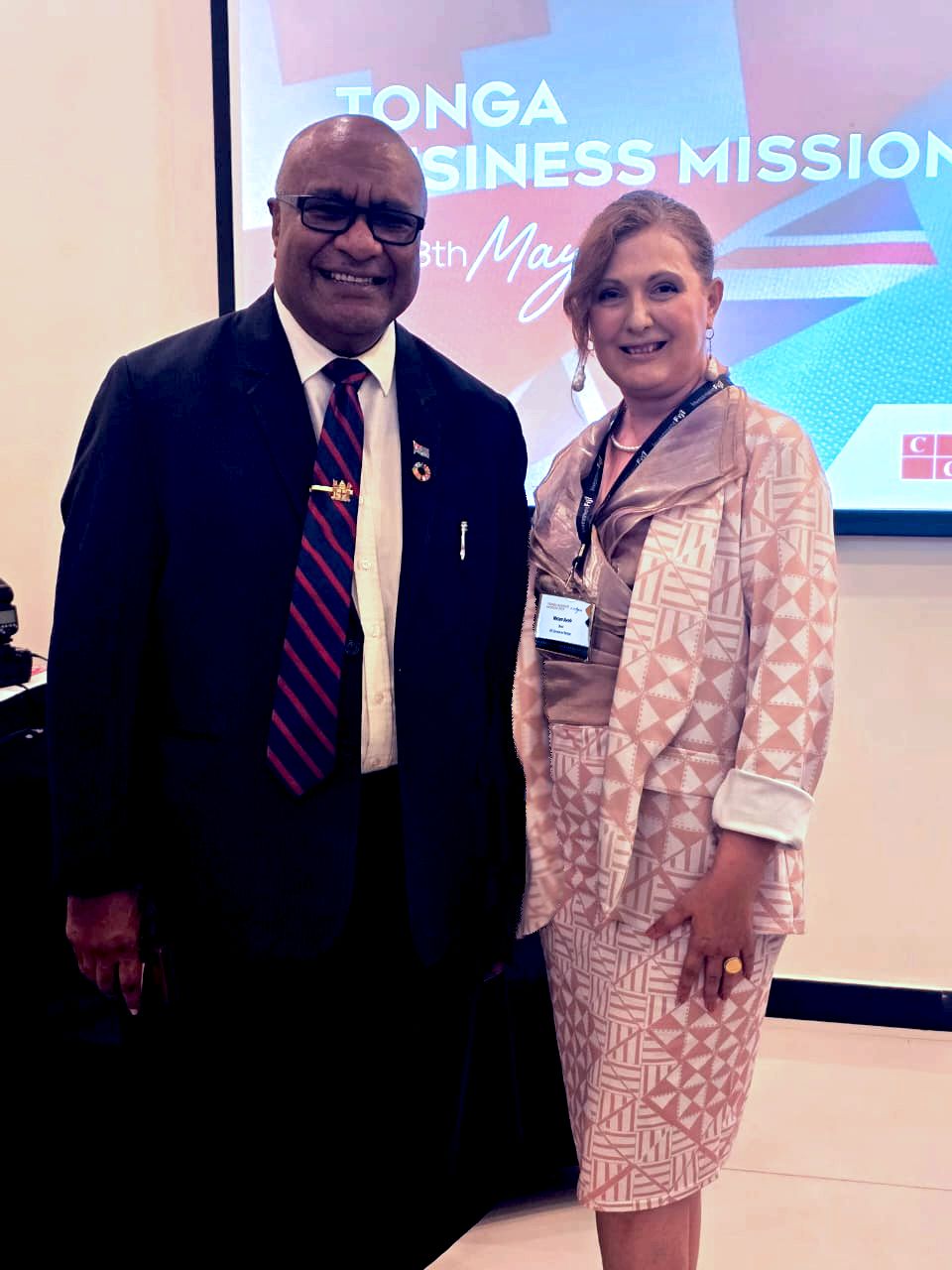 Two professionals posing together at the Tonga Business Mission event, with the woman wearing a patterned beige outfit and the man in a suit with a striped tie, both smiling in front of a colorful event backdrop.