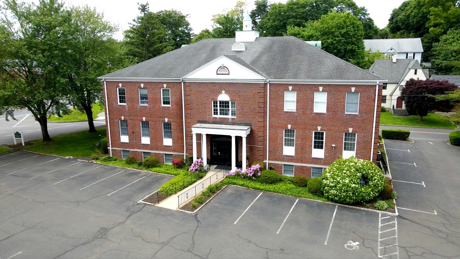 Two-story red brick commercial office building with white columns at the entrance, surrounded by landscaped greenery and a spacious parking lot, located in a suburban setting.