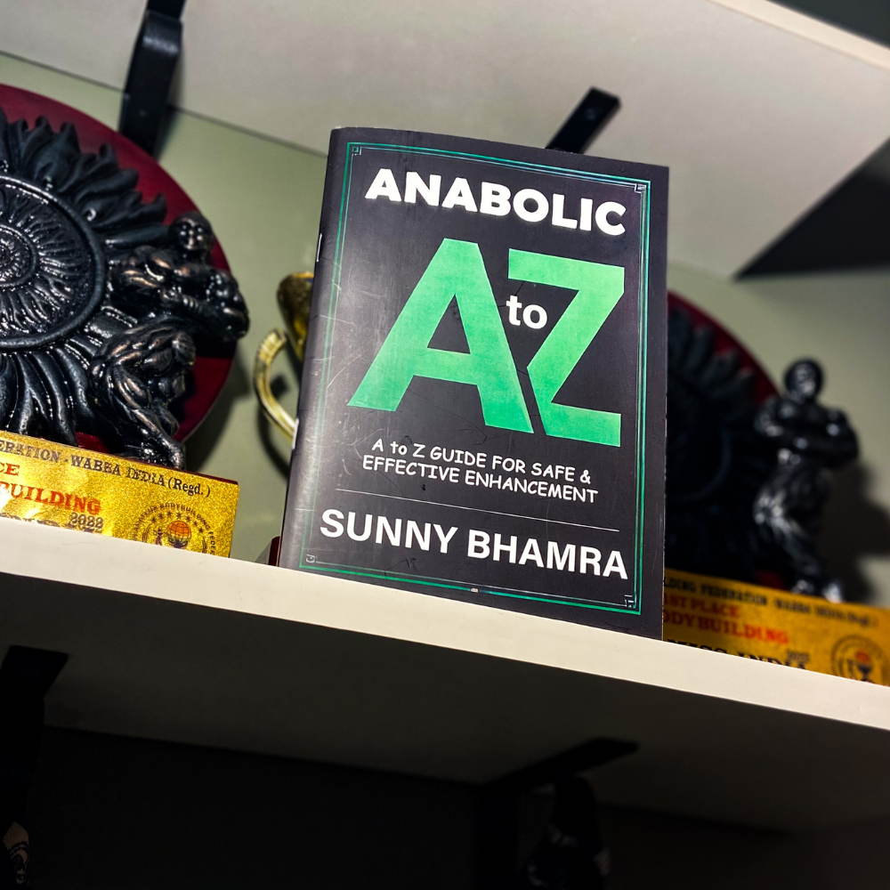 Close-up of the book 'Anabolic A to Z' by Sunny Bhamra, displayed on a shelf with bodybuilding trophies and a decorative item in the background, offering a guide for safe and effective enhancement.