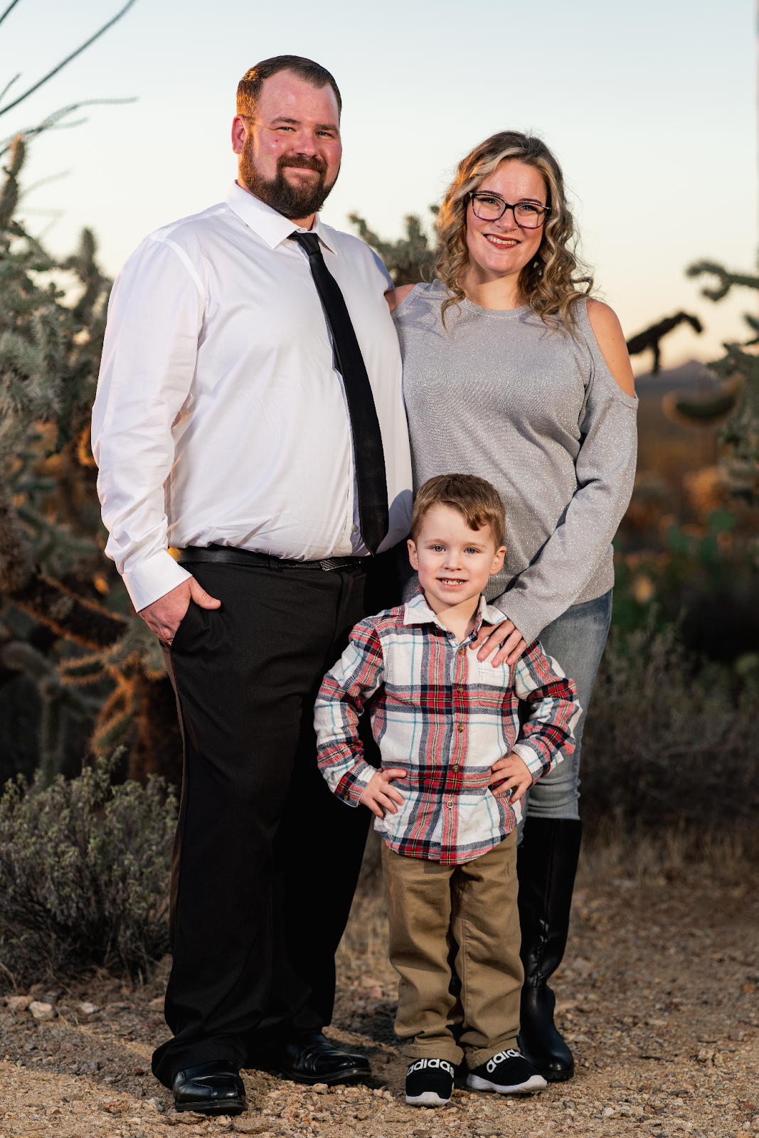 A family of three, including a father, mother, and young son, standing outdoors with a desert backdrop. The father is dressed in a white shirt and black tie, while the mother wears a silver sweater, and the son sports a plaid shirt and black sneakers. The family is smiling and posing together for a portrait.