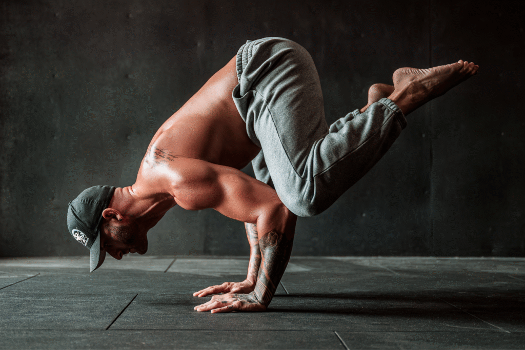 The man is performing a challenging bodyweight exercise, balancing on his hands in a gym setting. His body is in a plank position with his legs extended out.