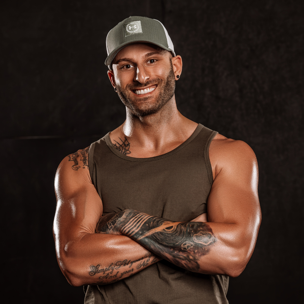 The man smiles at the camera, wearing a green cap and a brown tank top, with his arms crossed. The background is dark, and his muscular physique is highlighted by soft lighting.