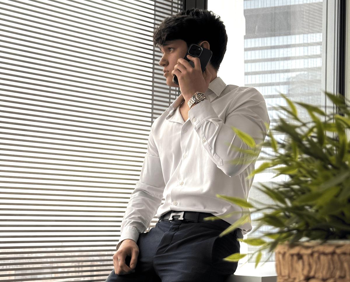 A young man in a white shirt and dark pants, standing by a window and talking on the phone while leaning against a desk.