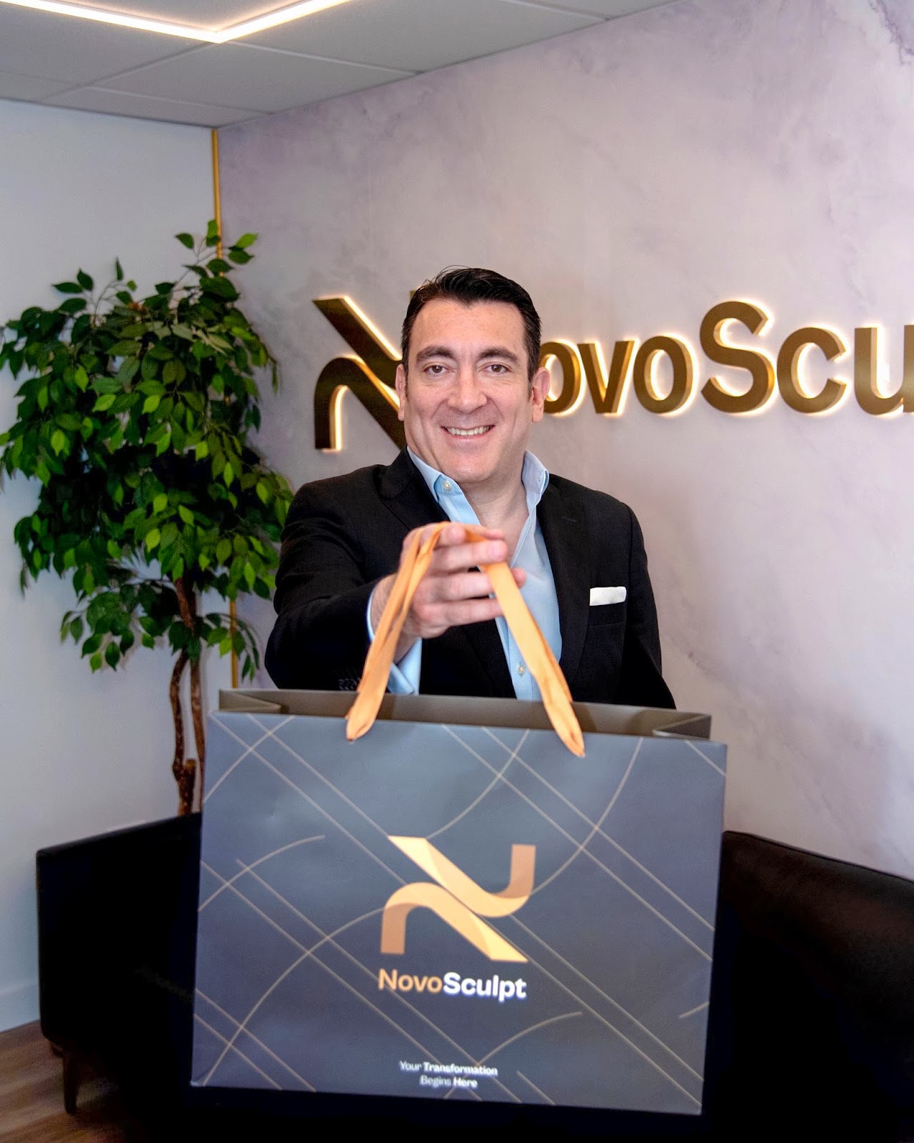 A man smiling and holding a NovoSculpt branded shopping bag, standing in front of the NovoSculpt logo on a marble wall. The image showcases the brand's commitment to transformation and innovation in fat removal technology.