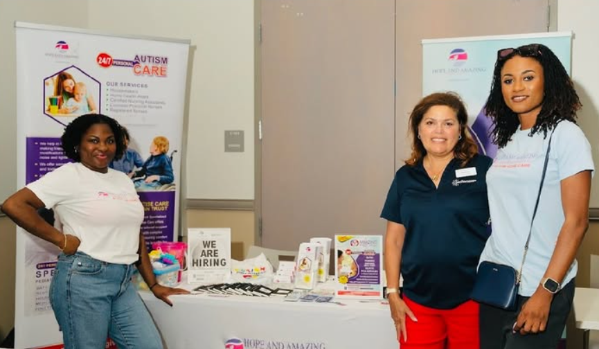 Two women standing at a promotional booth for 'Hope and Amazing Comfort Care LLC,' a company providing 24/7 personal autism care. One woman is smiling in a white t-shirt with the company logo, while the other wears a navy polo shirt. A table with brochures, business cards, and a 'WE ARE HIRING' sign is visible in the foreground.