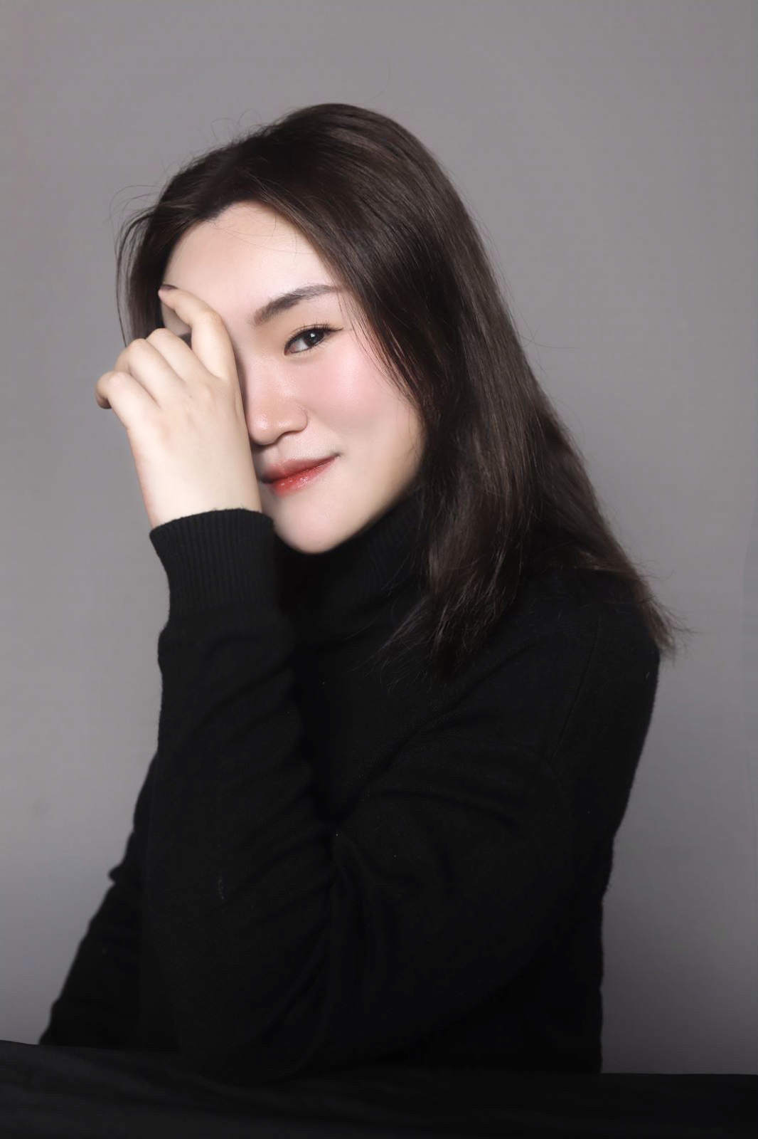 A close-up portrait of a woman in a black turtleneck sweater, smiling softly while holding one hand near her face, partially covering her eye. The background is neutral, adding to the calm, serene vibe of the image.
