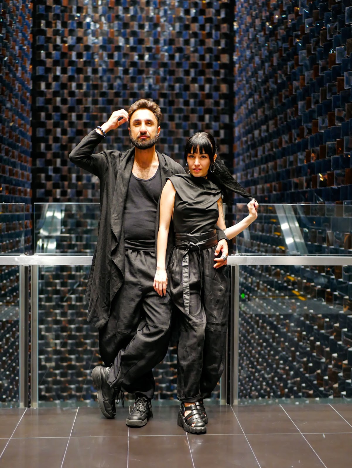 A stylish couple posing in black outfits against a backdrop of mirrored walls with a modern, geometric design.
