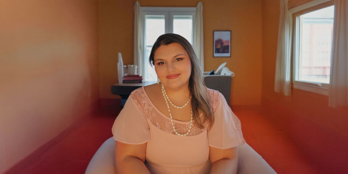 A woman with long hair wearing a pink blouse and pearl jewelry, sitting in a cozy room with orange walls and a window in the background. She is smiling gently with her hands resting on her lap."