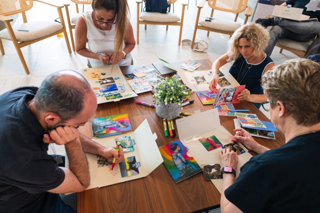 Several people seated at a table, working on creative projects with markers, stickers, and photographs.