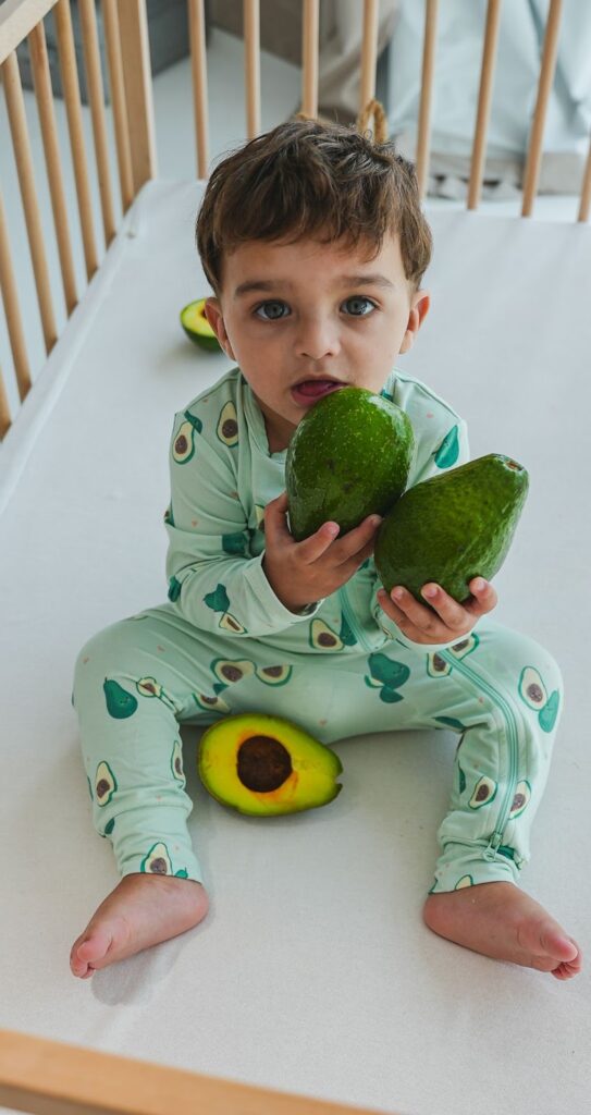 Child sitting in a crib holding two avocados, looking at the camera.