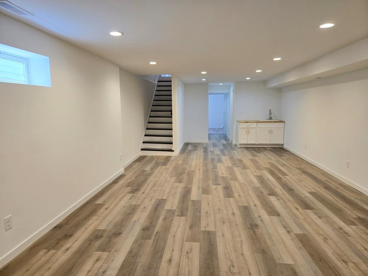 Spacious modern basement with light-colored wood flooring, recessed lighting, a small kitchenette area, and a staircase leading upstairs.
