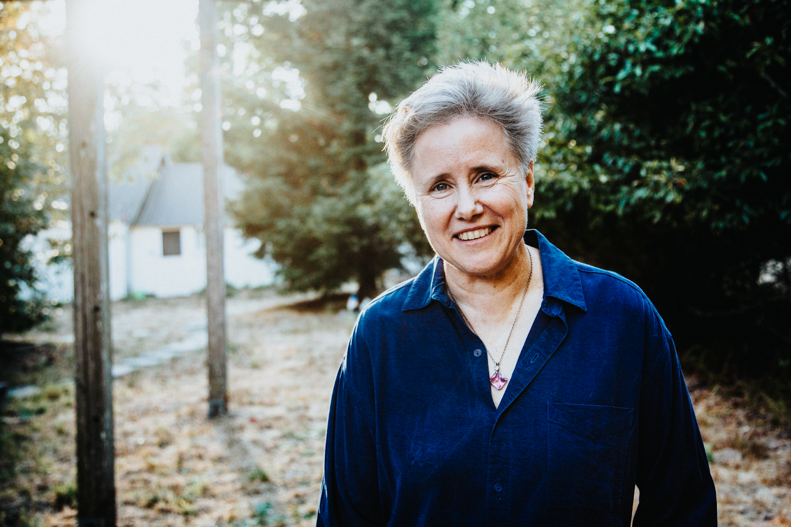 A smiling woman wearing a blue shirt, standing outdoors with sunlight shining through the trees behind her, creating a warm and inviting atmosphere.