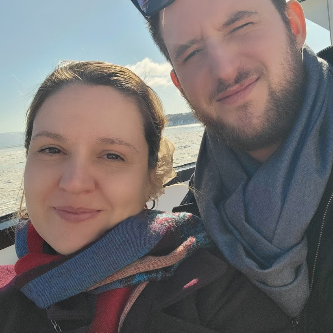 Couple enjoying a scenic boat ride on a sunny day, with the woman wearing a colorful scarf and the man dressed in a gray scarf.